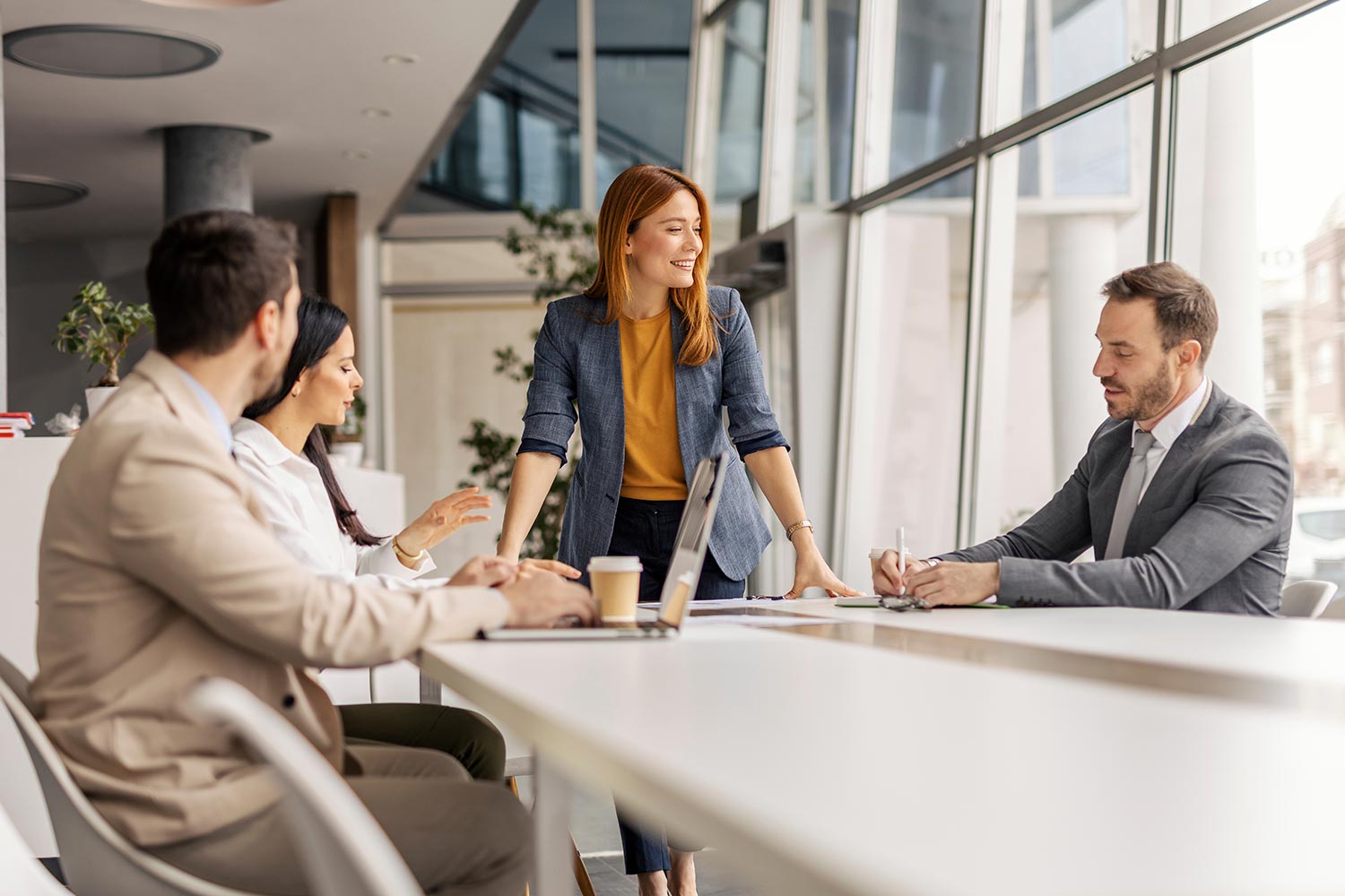 3 formally dressed people sat around a large table having a meeting, with a red-headed woman stood at the head of the table laughing
