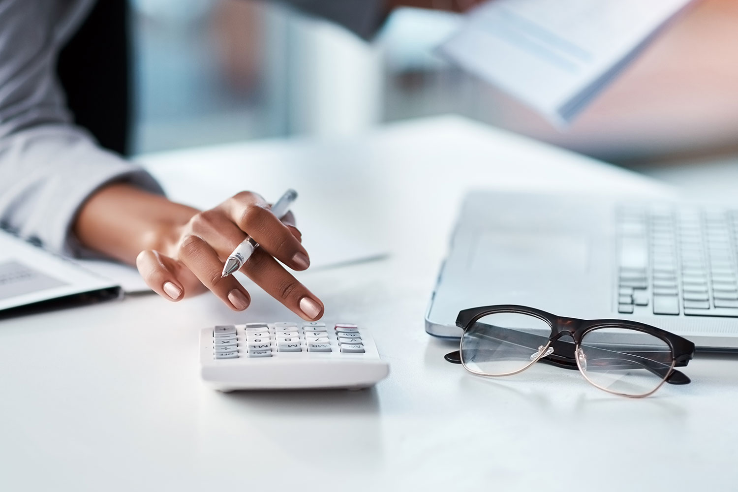 womans hand tapping on a calculator with pen in hand, with a pair of glasses resting on the table