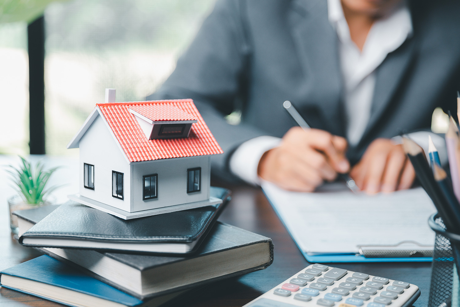 man in background writing on clipboard with stack of books with a model house on top