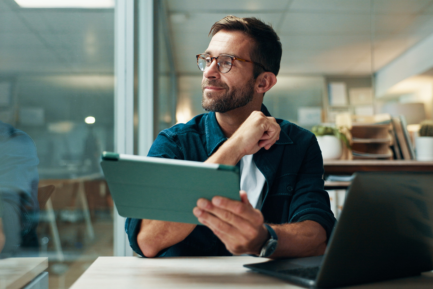 man wearing glasses looking to his right, smiling. He holds a tablet with a laptop also in front of him on the desk