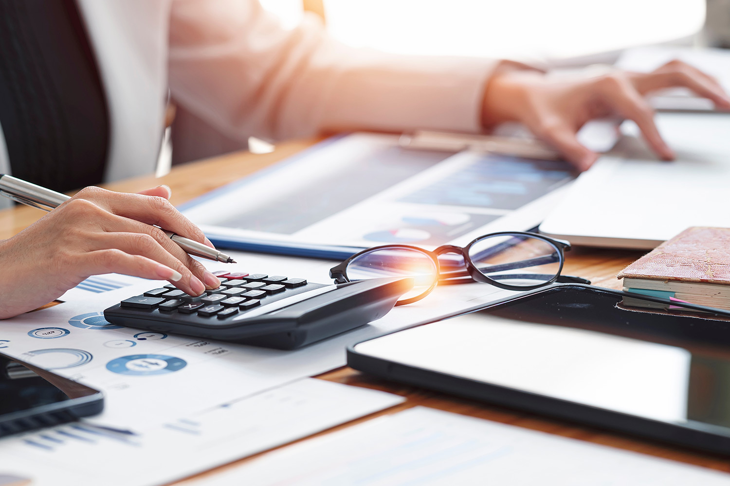 desk filled with miscellaneous work items such as glasses, papers, tablet. Woman uses calculator