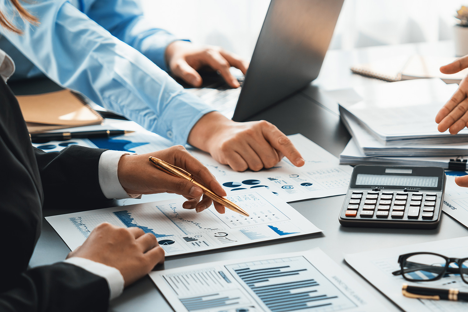 Multiple people discuss business matters, pointing to papers, calculators while someone works on a laptop