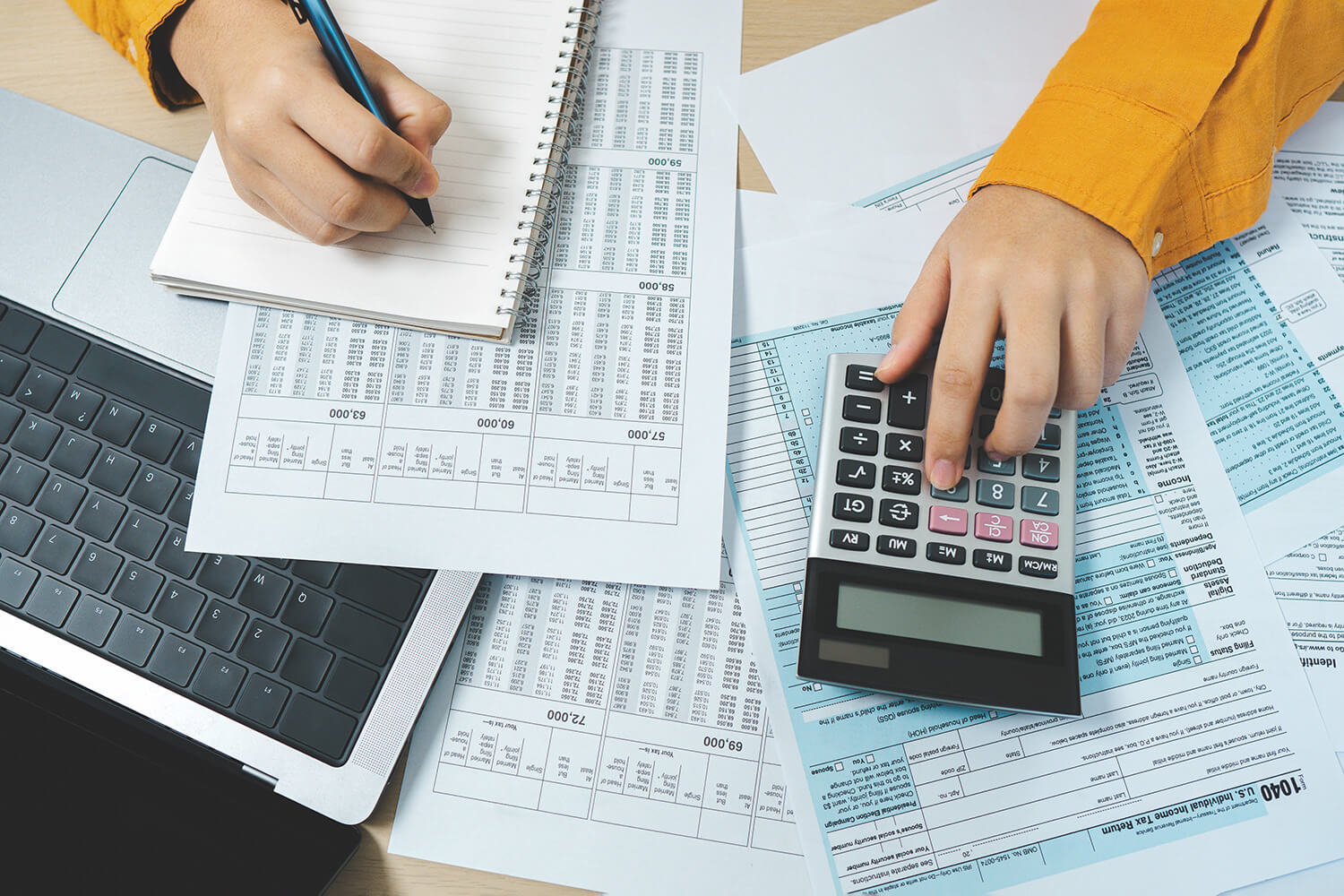 aerial shot of desk filled with accountancy documents, with a woman writing in a notebook with one hand and using a calculator with the other