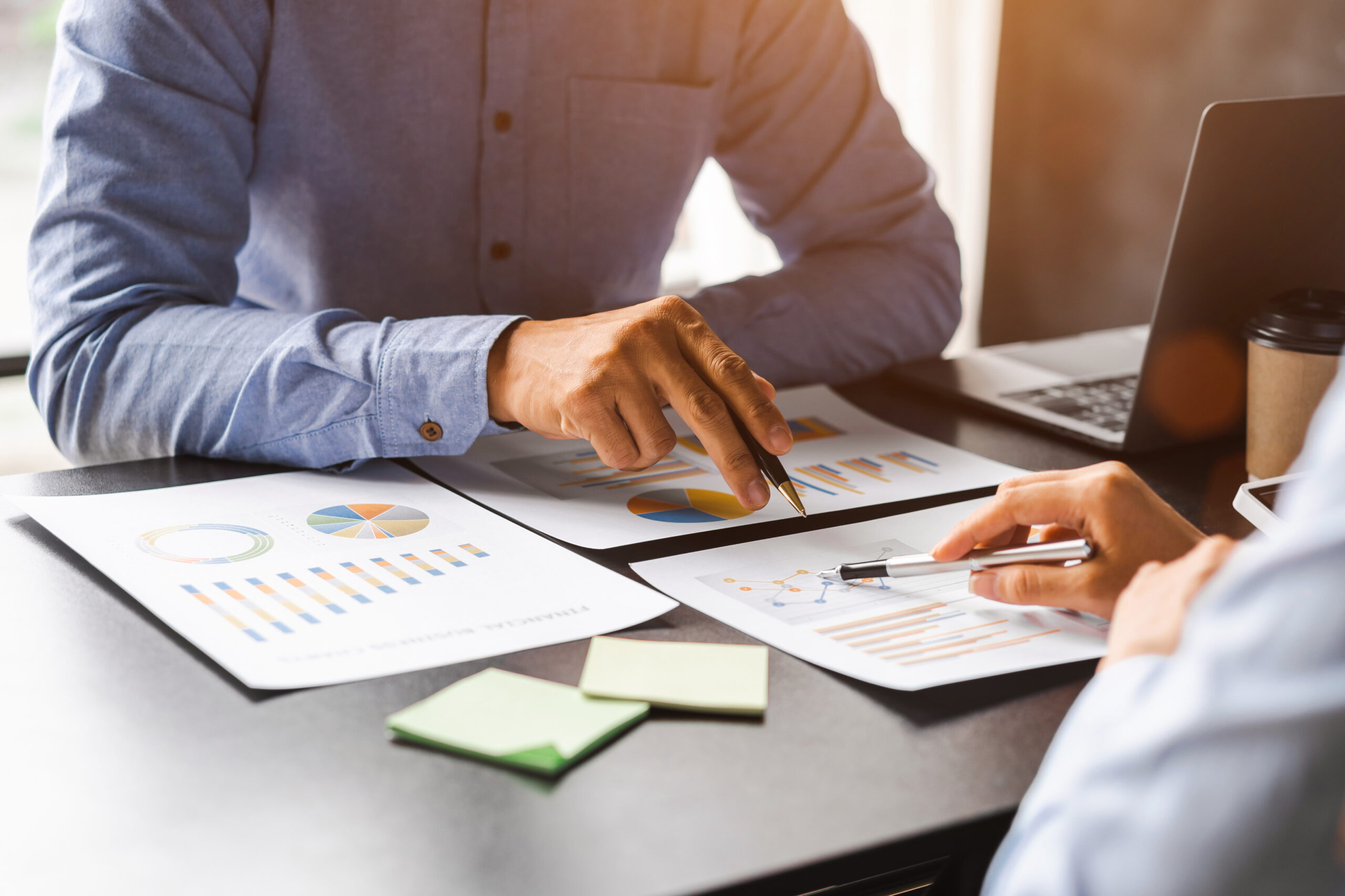 Desk with a man in shirt with a pen in his hand pointing at charts and grafts on the table to another person.