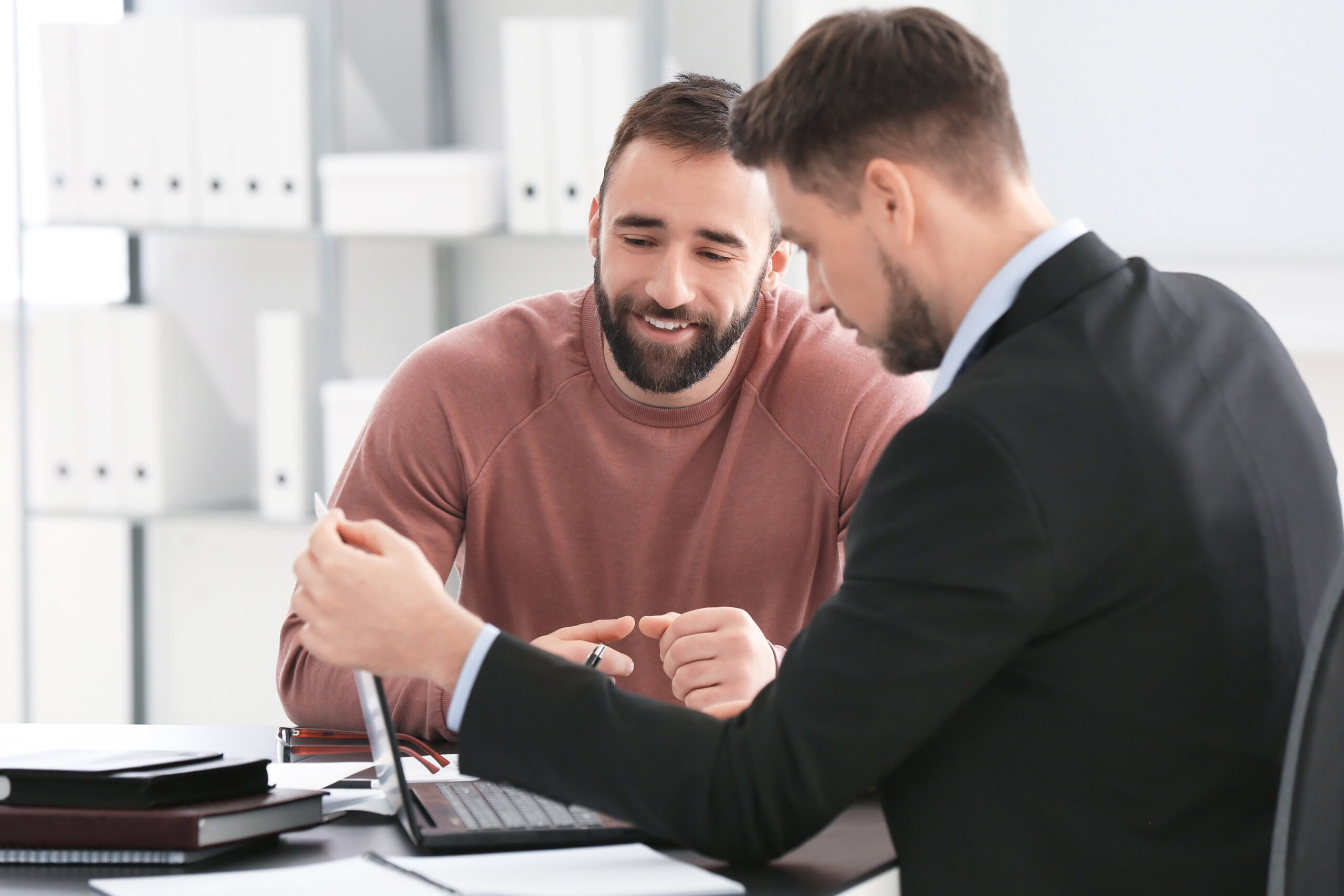A man in a suit explaining something on his laptop to a man in a red jumper with a beard in an office.