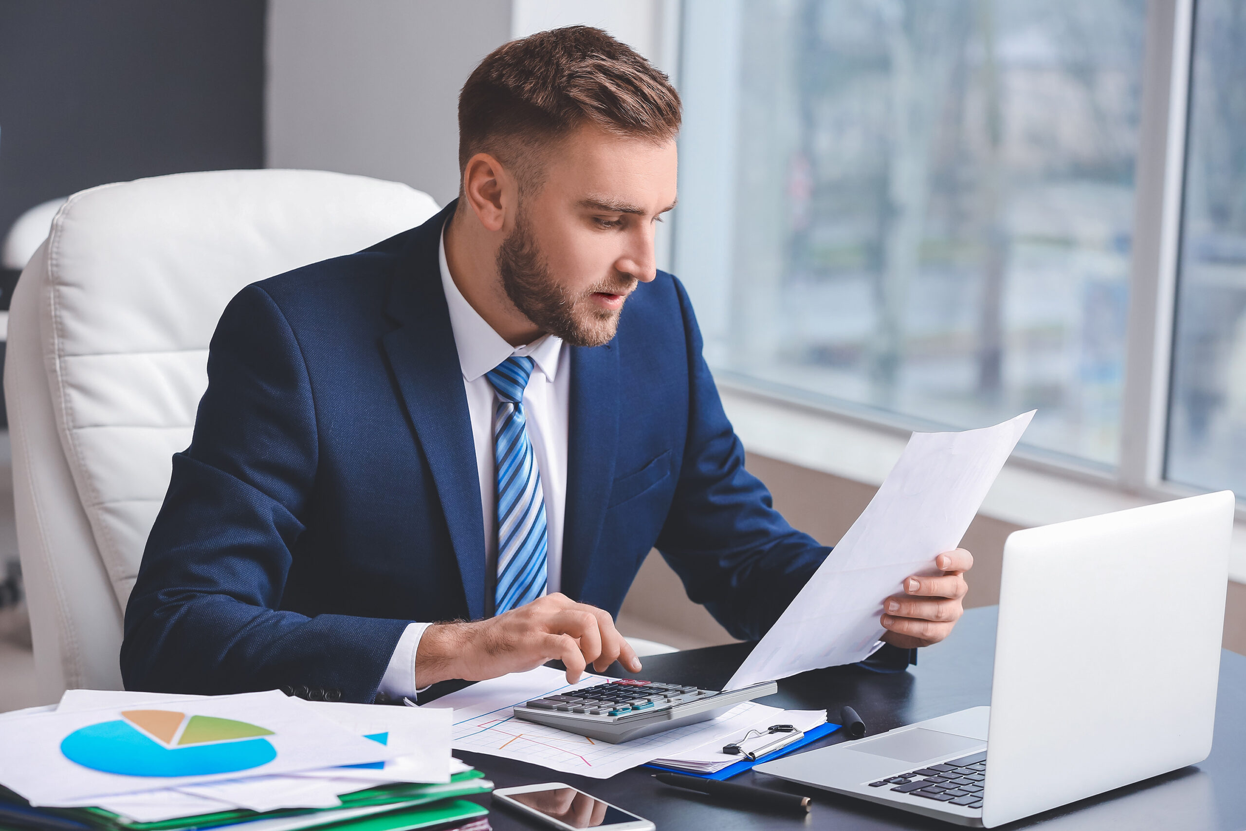 Man in a navy suit and tie reading some paperwork at his office desk using a calculator.